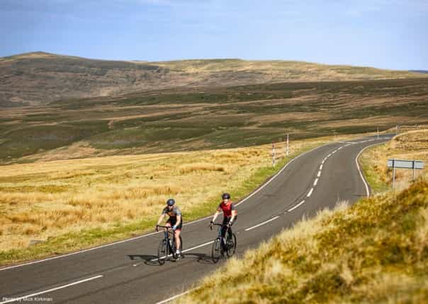 Two road cyclists riding a winding road through open moorland in the North Pennines