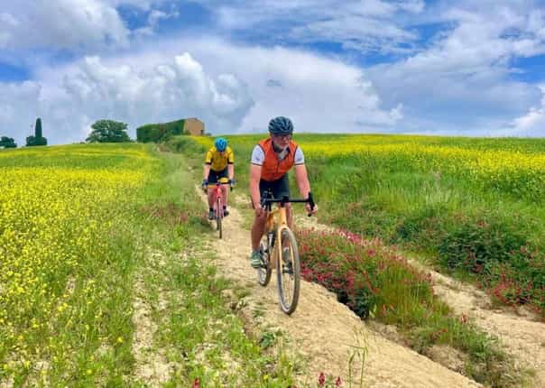 Two cyclists gravel riding on a narrow dirt track through vibrant yellow fields in Tuscany, Italy